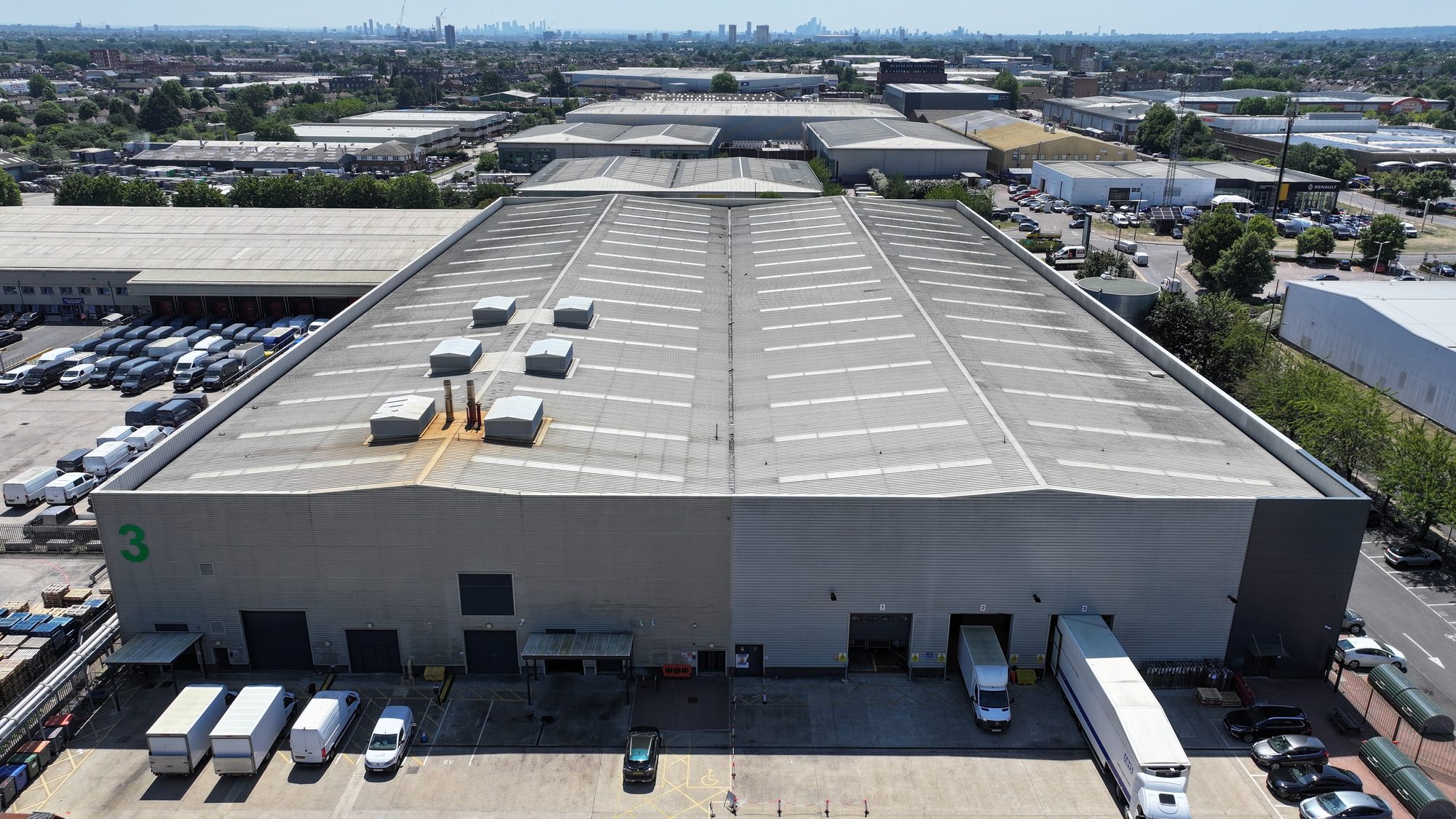 Aerial view of a large UK distribution warehouse with rooflight bands across a long span roof, HGVs at the loading bays