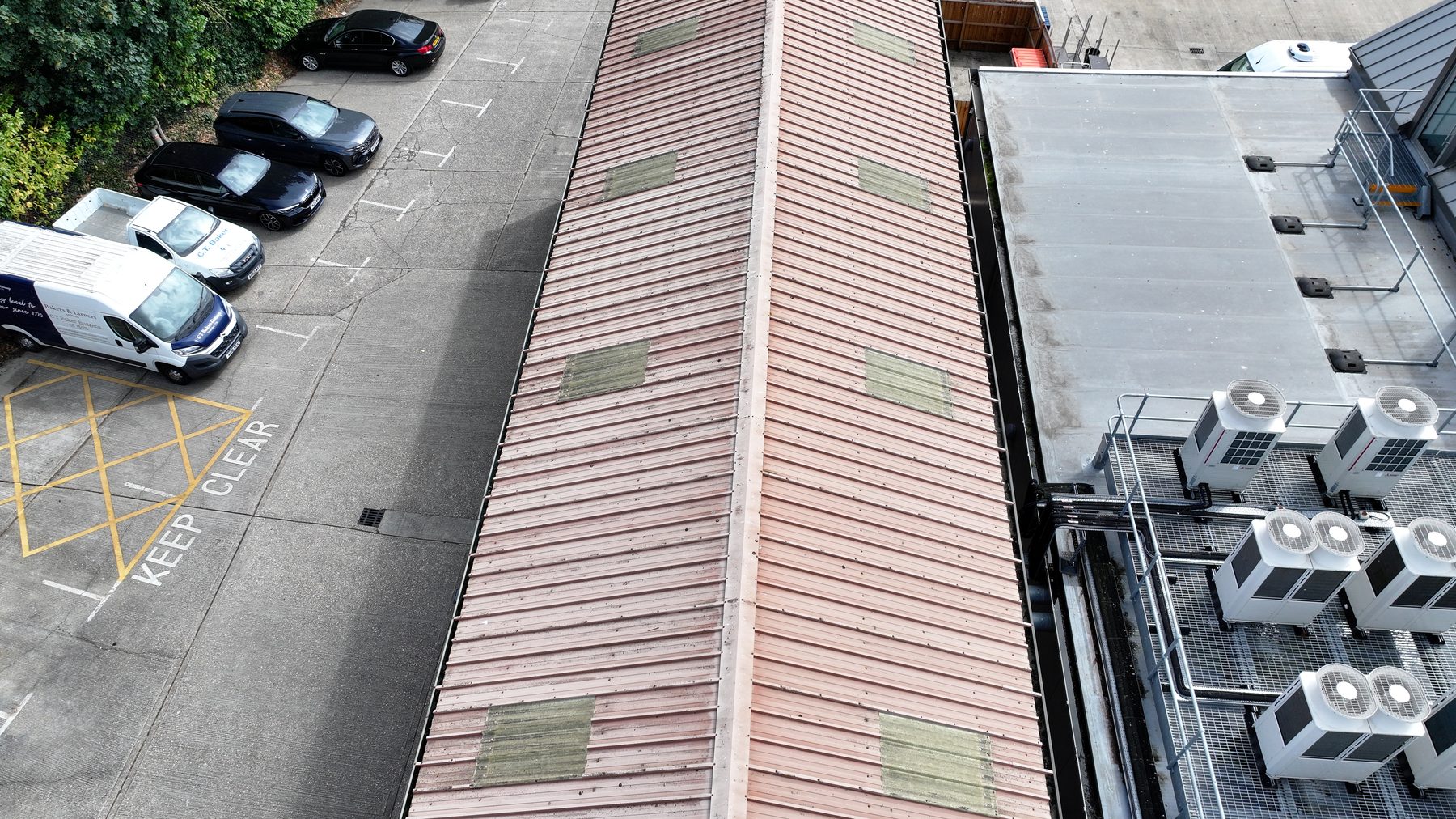 Aerial view of a pitched sheet roof beside a flat roof carrying a bank of air conditioning condenser units on a raised frame